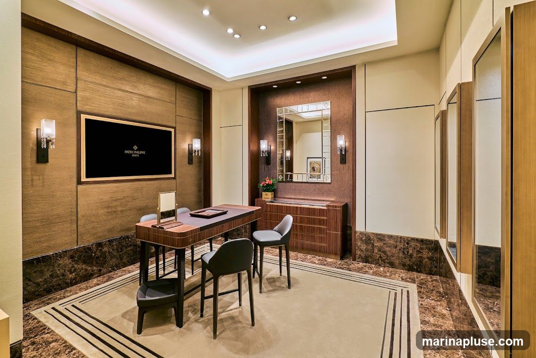 A luxurious showroom interior with a large screen displaying the Patek Philippe Geneva logo. A wooden table with chairs is set up for consultations, and mirrors adorn the walls. The space features rich wood paneling, marble accents, and elegant lighting.