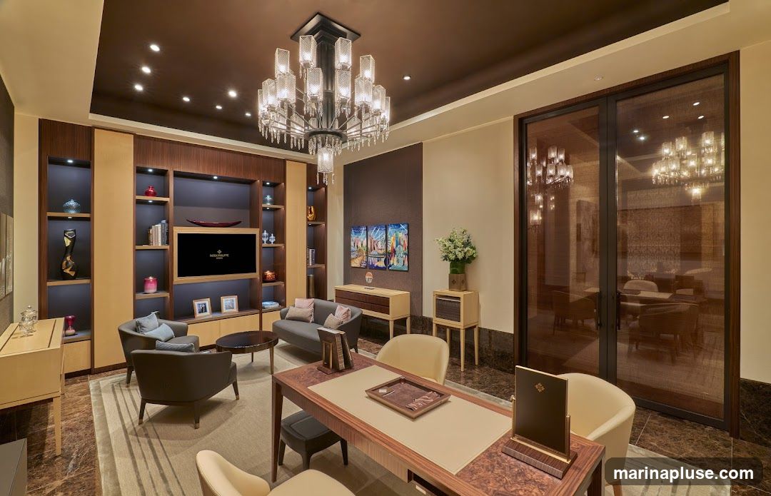 A well-lit, opulent office space featuring a large chandelier, plush seating, a television displaying a logo, and decorative shelves. The room is furnished with a desk, chairs, a sofa, and a coffee table, creating a sophisticated and comfortable atmosphere. The walls are adorned with artwork, and the flooring is a rich marble.