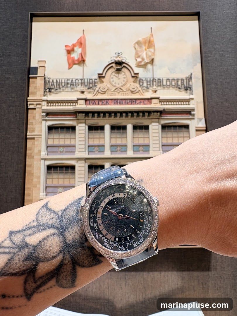 A close-up of a Patek Philippe watch on a wrist, with a miniature diorama of the Patek Philippe manufacture building in the background. The watch face displays world time zones. The diorama features Swiss and orange flags, a clock, and the text "MANUFACTURE O HORLOGERIE" and "PATEK PHILIPPE & CO.". A tattoo of a flower is visible on the wrist.