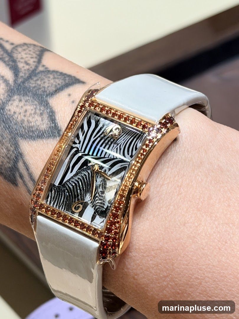 A close-up shot of a woman's wrist wearing a rectangular watch with a white band. The watch face features a black and white zebra pattern with a gold accent. The bezel of the watch is adorned with small red stones. The woman has a tattoo on her arm.