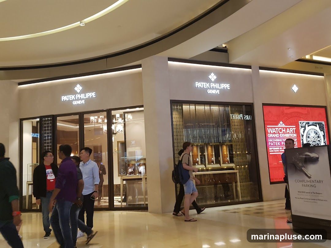 The exterior of a Patek Philippe store with its name and logo prominently displayed above the entrance. The store features large glass windows showcasing watches and display cases. To the right, a large red advertisement for a "Watch Art Grand Exhibition" is visible, along with a sign for "Complimentary Parking." Several people are walking past the storefront.