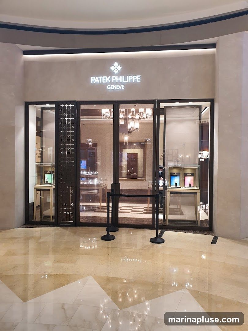 The exterior of a Patek Philippe Geneve watch store. The store has a large glass facade with illuminated signage above that reads "PATEK PHILIPPE GENEVE". Inside, display cases showcase watches. Black stanchions with ropes are placed in front of the entrance. The floor is polished marble.