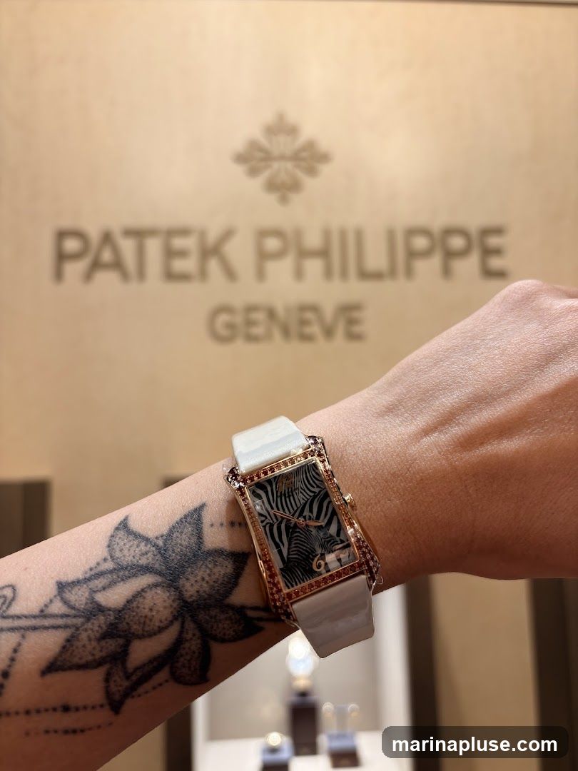 A close-up of a person's wrist wearing a Patek Philippe watch with a zebra print face. The watch has a rose gold case and a white strap, adorned with small red stones. The background features the Patek Philippe Geneve logo embossed on a textured wall. Below the logo, display cases with watches are visible. The person has a large lotus flower tattoo on their arm.