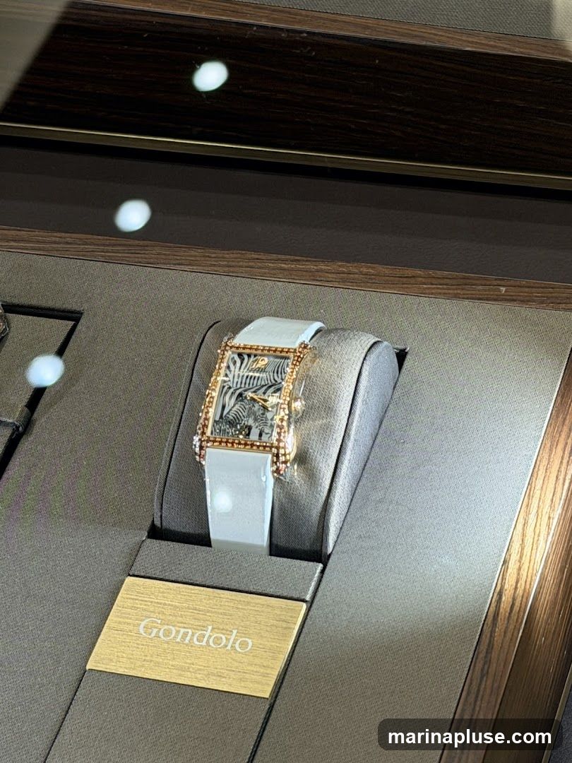 A close-up view of a luxury watch with a zebra pattern on its face, displayed in a velvet-lined case. The watch has a rectangular gold case with diamond accents and a light blue leather strap. A golden plaque below the watch reads "Gondolo". The display case has a dark wood finish.
