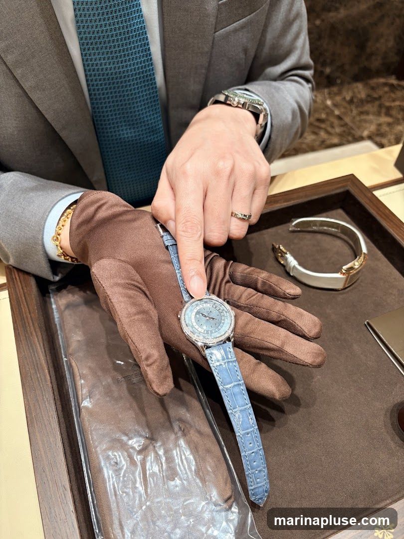 A person wearing a suit, tie, and gloves is presenting a luxury watch with a light blue alligator strap. The watch face is intricate and appears to be adorned with small diamonds. Another watch band is visible in the background. The presentation is taking place on a dark brown display tray, suggesting a retail or exhibition setting.