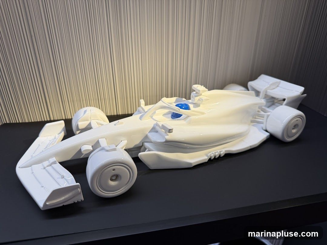 A glossy white model of a Formula 1 race car is displayed on a black shelf against a textured gray background. The model features intricate details, including the front and rear wings, wheels, and a blue cockpit.