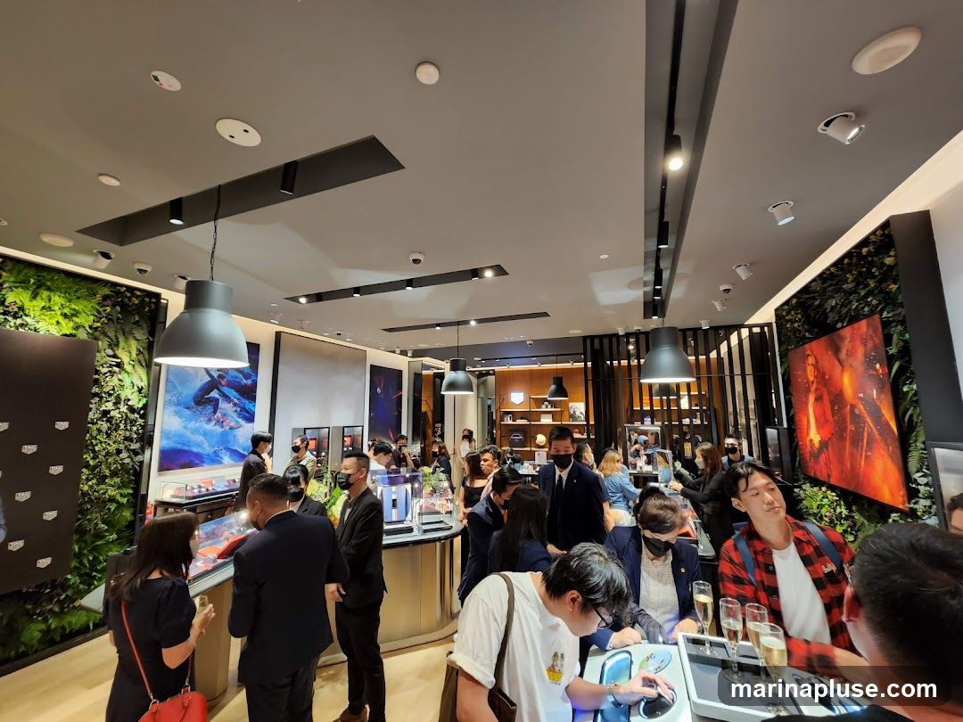 A group of people are gathered inside a well-lit store, with some looking at displays and others conversing. The store features modern decor, including green wall accents, large artwork, and prominent lighting fixtures. Several individuals are wearing masks.