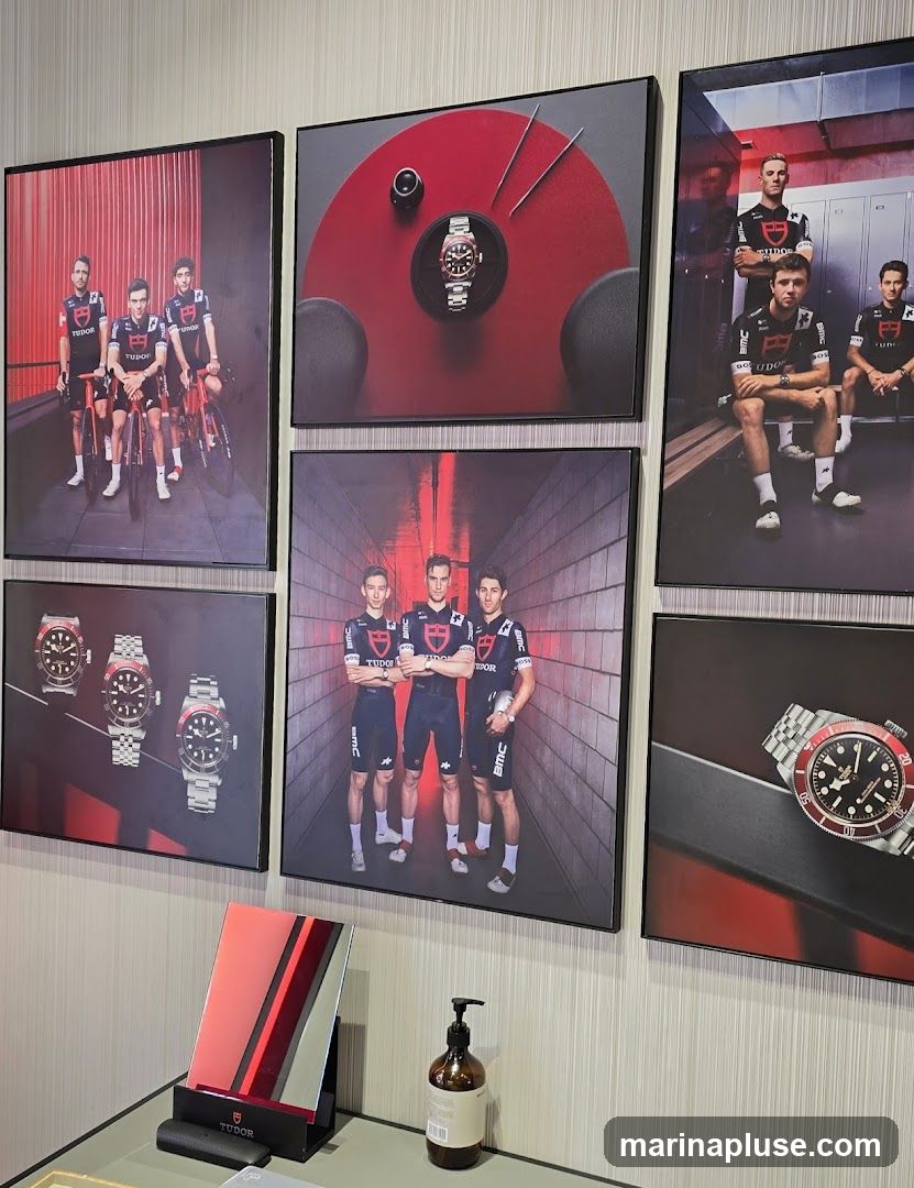 A wall display featuring framed photographs of Tudor watches and athletes wearing Tudor branded cycling attire. The photographs showcase different watch models and group shots of cyclists. In the foreground, a small mirror with a Tudor branded stand and a bottle of soap are visible on a counter.