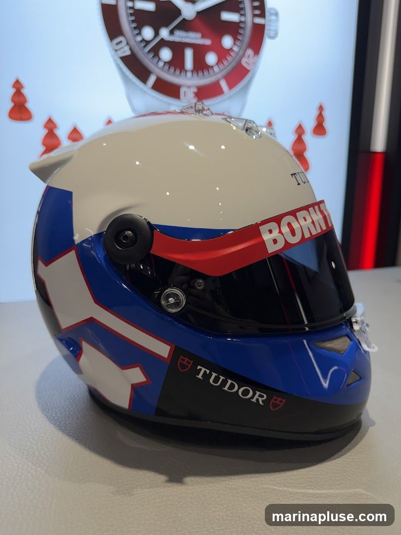 A white, blue, and red racing helmet with the TUDOR logo is displayed on a white surface. Behind the helmet, a large watch face with a red dial is visible, along with several small red tree-like decorations. The word "BORN" is visible on a red band above the helmet's visor.