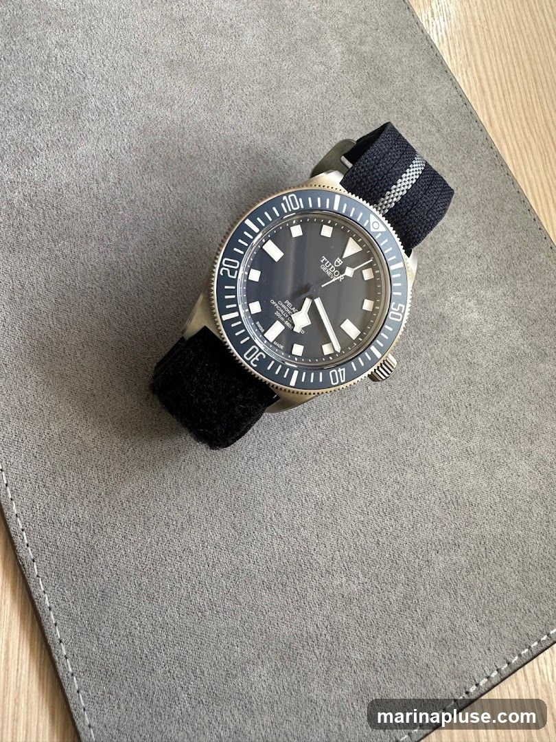 A close-up shot of a black Tudor Pelagos dive watch with a black fabric strap. The watch is placed on a grey textured mat, with a wooden surface visible in the background. The watch face shows white hour markers and hands, with the Tudor logo and "Pelagos" text clearly visible. The bezel is marked with white numerals and indices.
