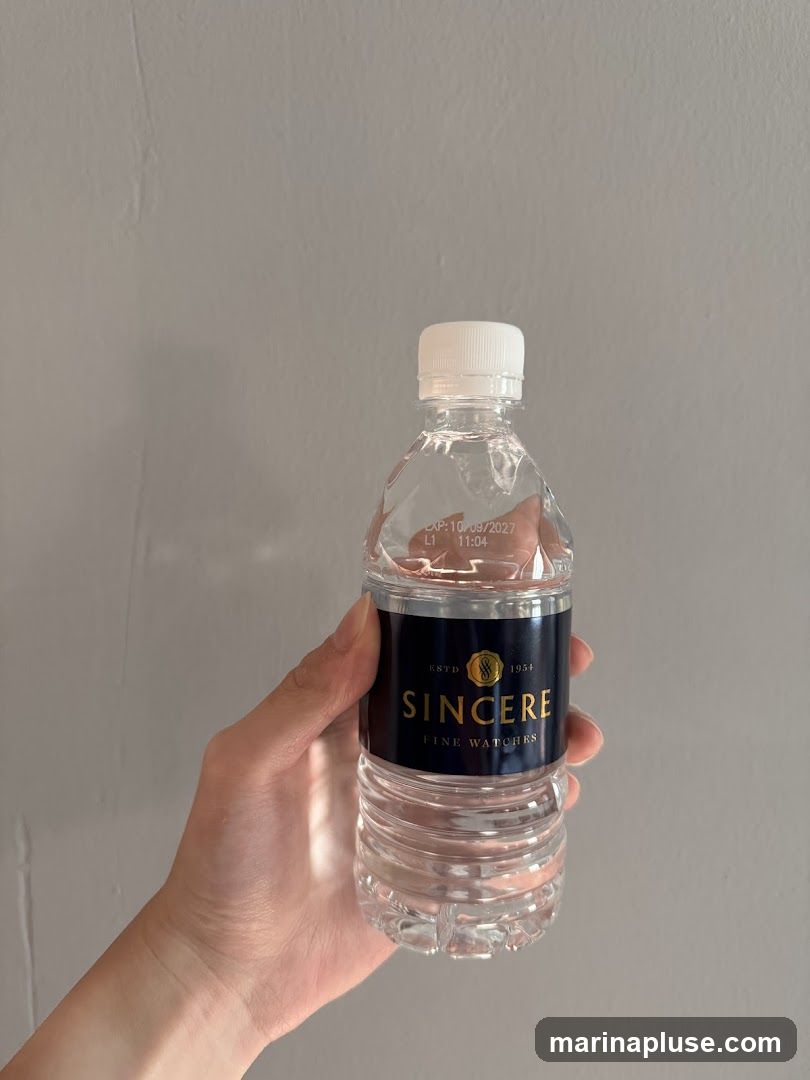 A hand holds a clear plastic bottle of water with a black label. The label has gold text that reads "SINCERE FINE WATCHES" and "ESTD 1954". There is also an expiration date printed on the bottle.