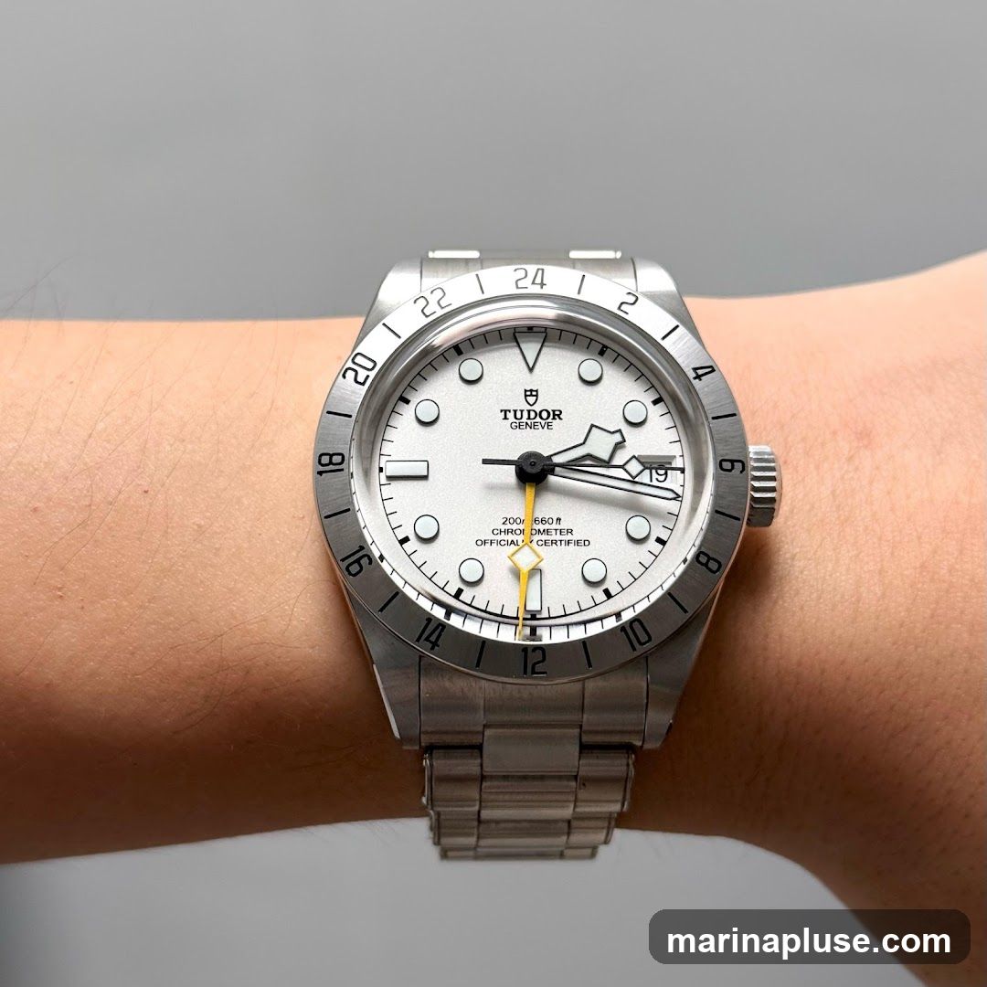 A close-up shot of a silver Tudor watch with a white dial, worn on a person's wrist. The watch features a GMT bezel with 24-hour markings, a date window at the 3 o'clock position, and a yellow GMT hand. The dial has circular hour markers and luminous hands. The watch is attached to a stainless steel bracelet.