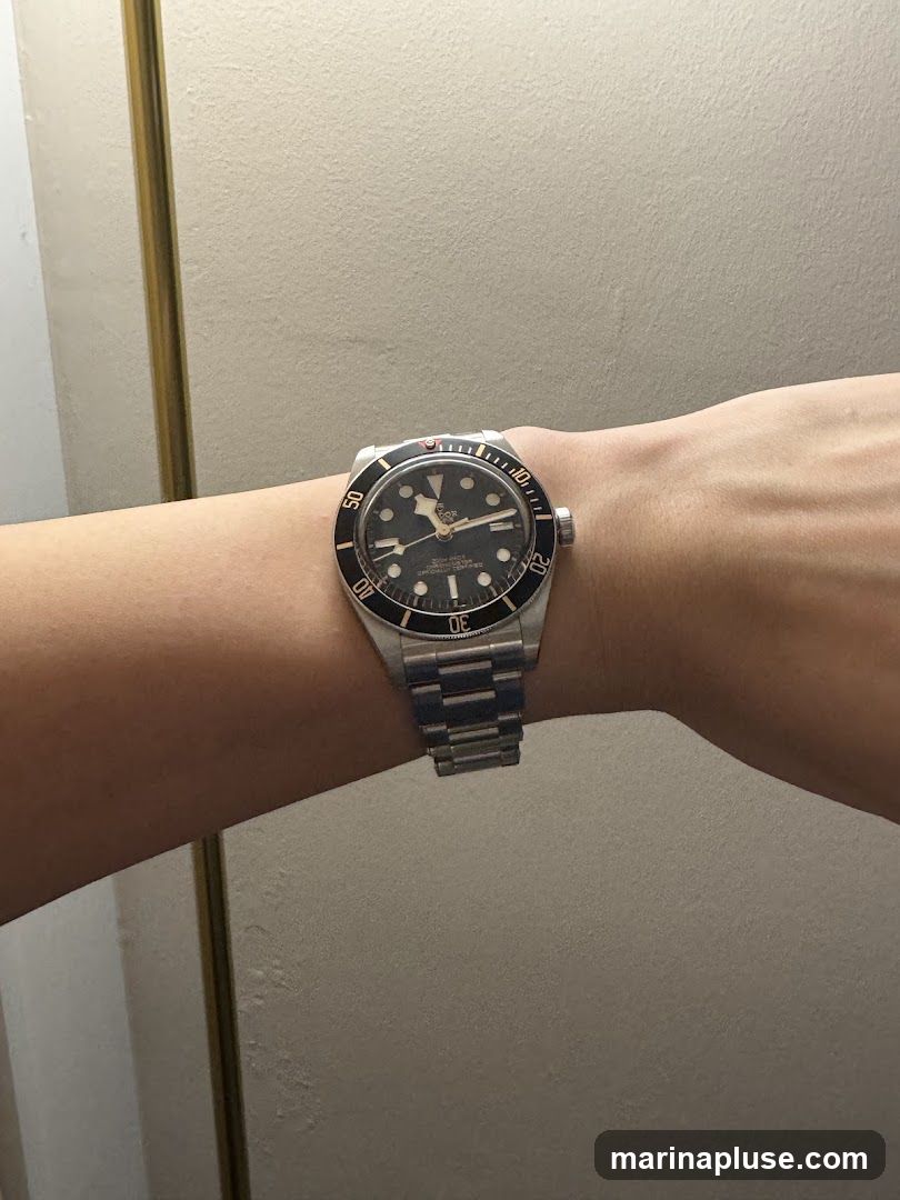 A close-up shot of a person's wrist wearing a stainless steel watch with a black dial and bezel. The watch has a metal link band. The background is a textured, light-colored wall.