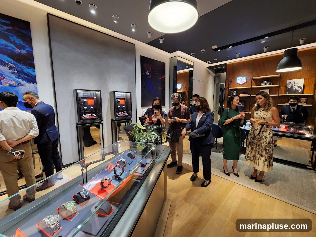 A group of people are gathered inside a modern watch store, looking at displays of watches. The store features sleek display cases, artwork on the walls, and shelves stocked with merchandise. A prominent TAG Heuer logo is visible on the back wall.