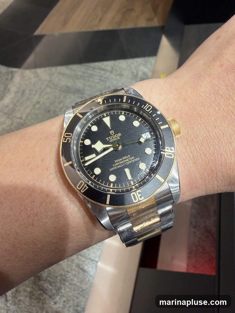 A close-up shot of a Tudor watch on a person's wrist. The watch has a black dial with gold accents and a two-tone metal band. The bezel is black with gold markings. The watch appears to be new, with protective plastic still on the band.