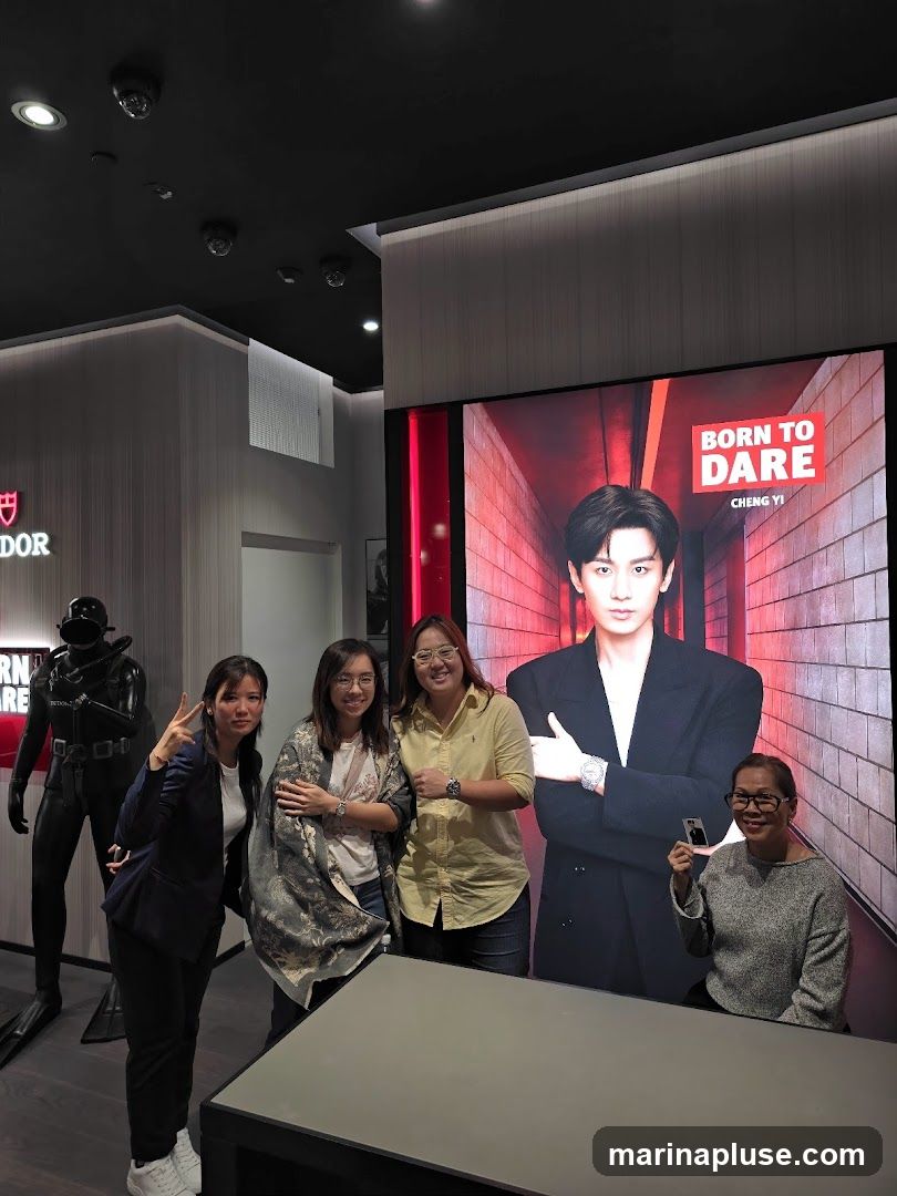 Four women are posing for a photo in what appears to be a retail store. To the left, a mannequin dressed as a diver stands next to a display with the word "DOR" visible. In the background, a large screen displays a portrait of a man with the text "BORN TO DARE CHENG YI". The women are smiling and interacting with the display.