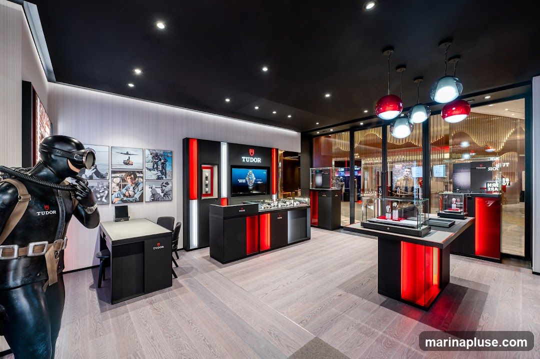 The interior of a Tudor watch store, featuring a life-size diver mannequin in the foreground. The store has a modern design with black and red accents. Display cases showcase various Tudor watches. The walls are adorned with images related to Tudor's history and diving. A large screen displays Tudor branding. The flooring is light-colored wood.