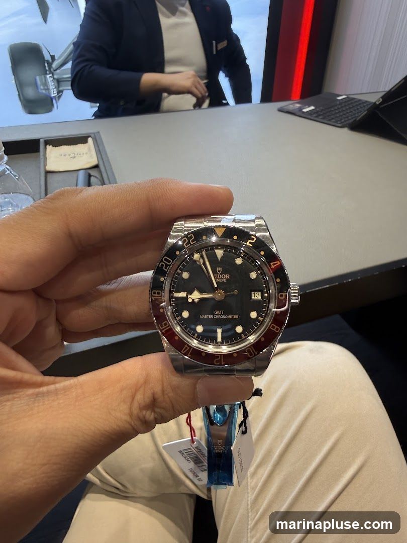 A person is holding a Tudor GMT Master Chronometer watch. The watch has a black dial with gold accents and a red and black bezel. The watch is new and has tags attached. In the background, a person in a suit is visible, possibly a salesperson, and a screen displaying an image of an airplane wheel.