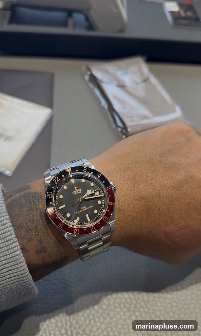 A close-up shot of a Tudor GMT watch on a person's wrist. The watch has a dark grey dial with a red and black bezel. The bracelet is made of stainless steel. In the background, there are some blurred items, possibly from a retail environment.