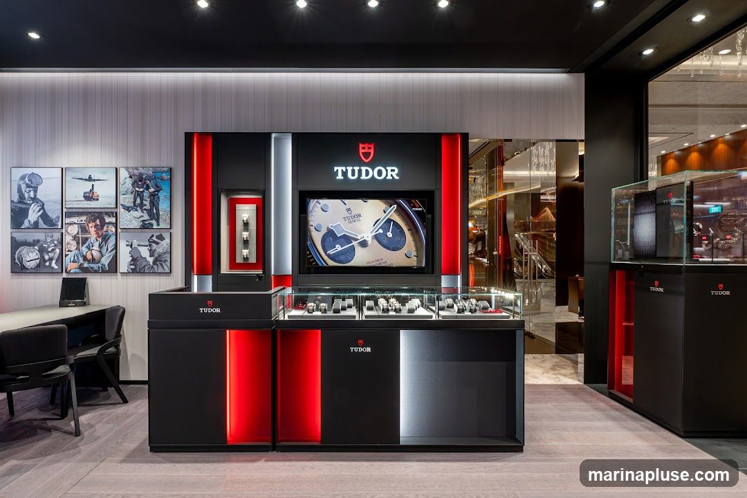A modern and sleek interior display for Tudor watches. The display features a large screen showing a close-up of a watch face, with the Tudor logo prominently displayed above. Below the screen, glass cases showcase an assortment of watches. To the left, a wall is adorned with framed black and white photographs depicting historical aviation and diving scenes. The display units are black with striking red and white accents.