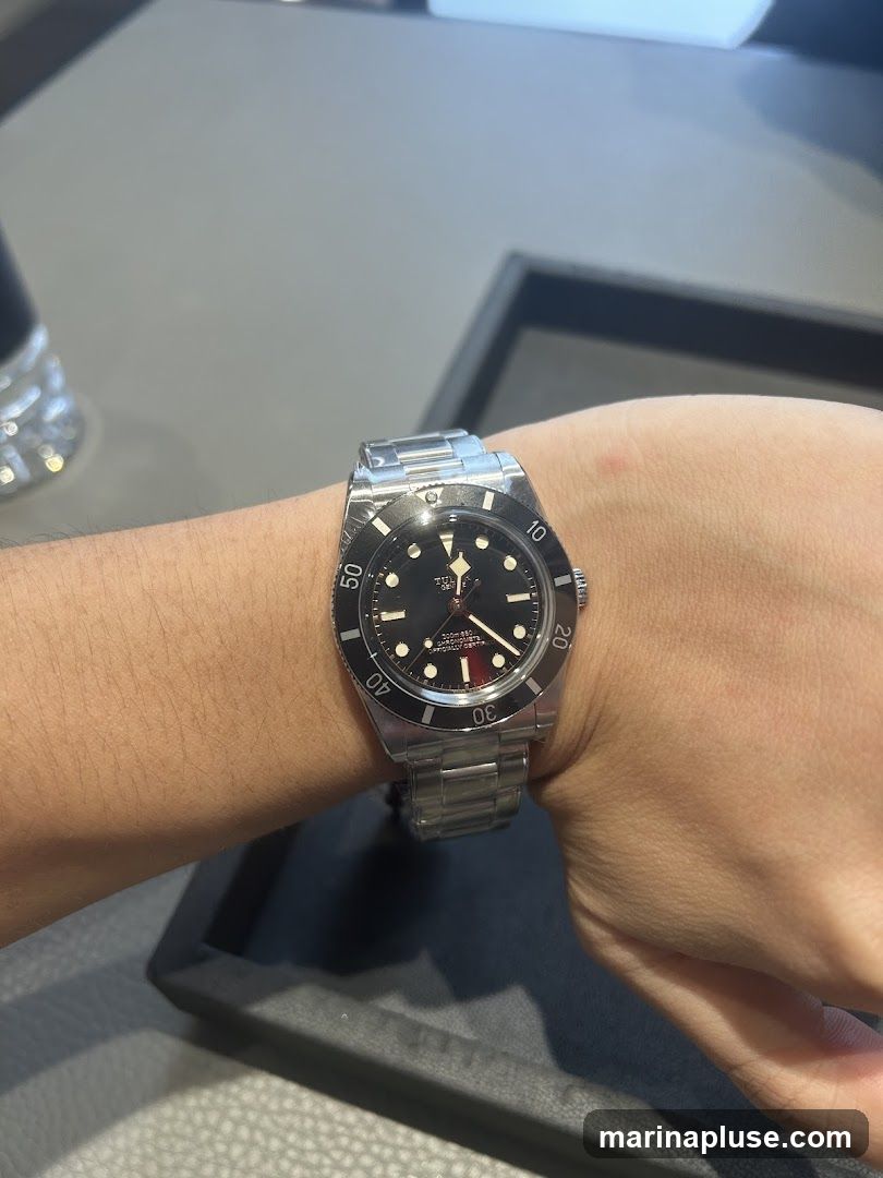 A close-up shot of a person's wrist wearing a black-faced wristwatch with a metal band. The watch has a rotating bezel with minute markers. The dial has white hour markers and hands, with a red accent at the 12 o'clock position. Text on the dial indicates "TUDOR" and "200m: 660ft". The background appears to be a display surface in a retail environment.