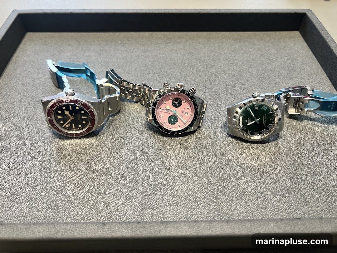Three luxury watches are displayed on a textured gray surface. The watch on the left has a black dial with a red bezel. The middle watch has a pink dial with chronographs. The watch on the right has a green dial with Roman numerals. All watches have metal bracelets.