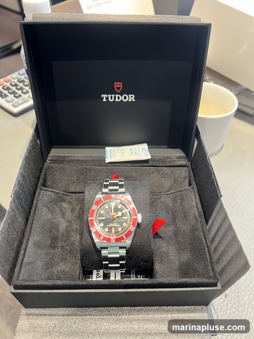 A Tudor watch with a black dial and red bezel is displayed inside its black presentation box. The Tudor logo is visible on the inside lid of the box. A silica gel packet is also present in the box. In the background, a calculator and a white mug are partially visible.