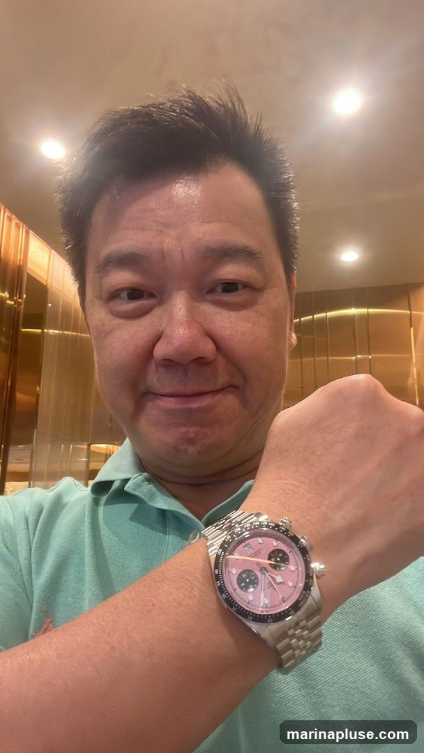 A man in a light green polo shirt is taking a selfie. He is wearing a silver watch with a pink face and black accents on his left wrist. The background appears to be an interior with gold-colored paneling and lighting.