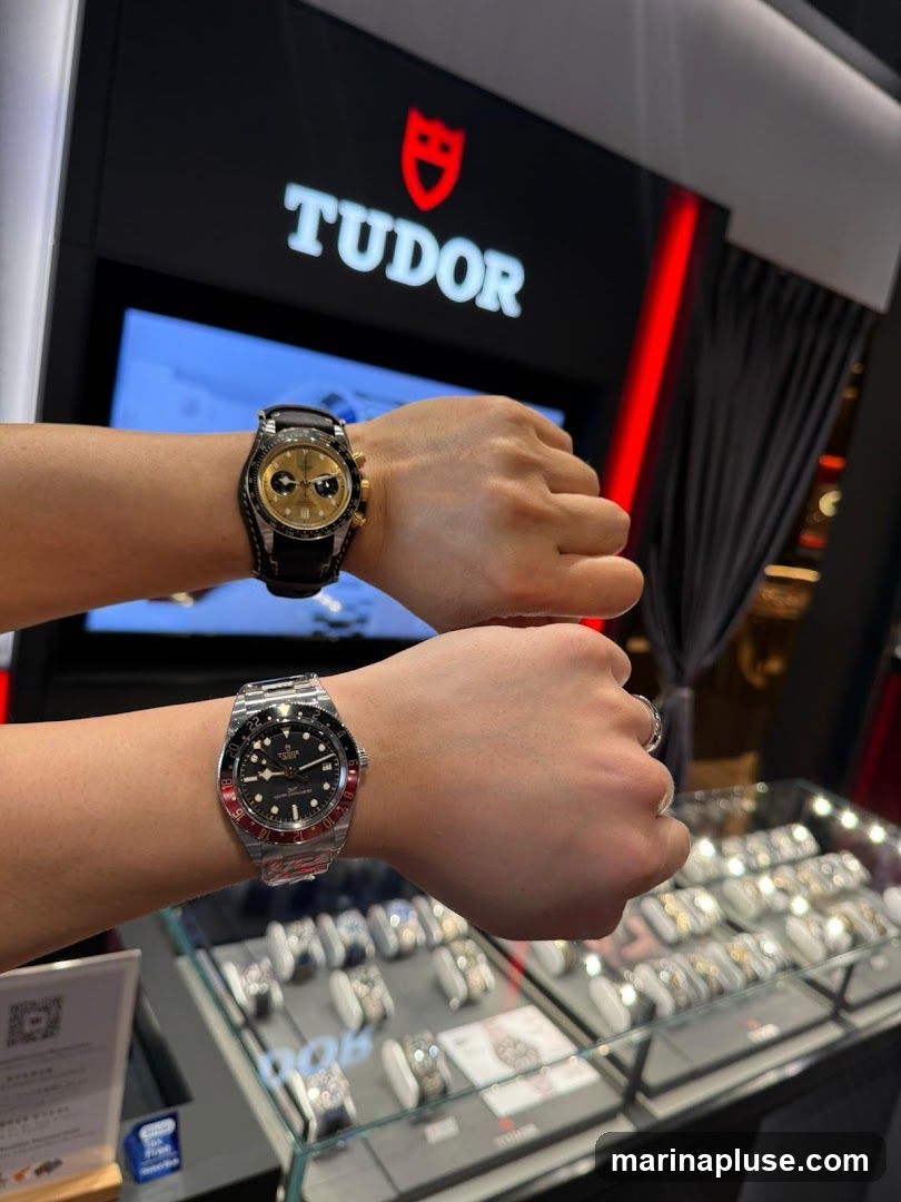 Two wrists are shown, each wearing a Tudor watch. The top wrist wears a gold-toned chronograph watch with a black leather strap. The bottom wrist wears a stainless steel watch with a black and red bezel. In the background, the TUDOR logo is prominently displayed in white and red on a black panel. Below the logo, a screen displays a blue image. A display case filled with watches is visible in the foreground.