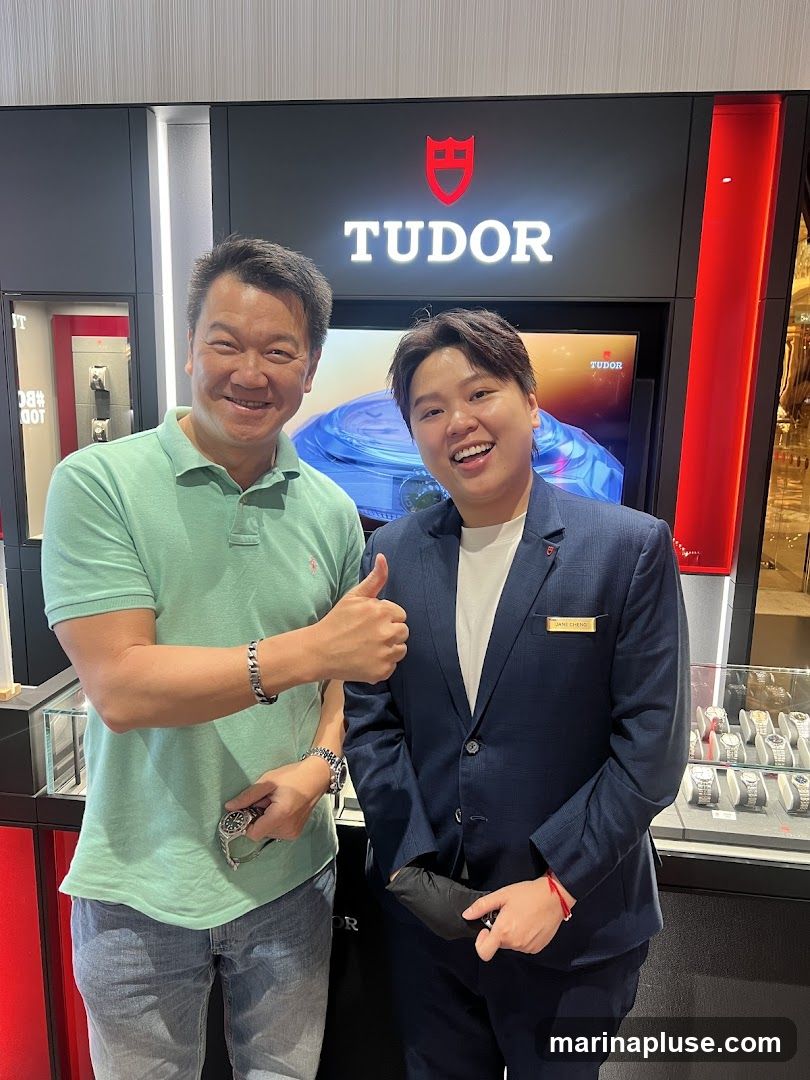 Two people are posing for a photo in front of a Tudor store display. The person on the left is wearing a light green polo shirt and jeans, and is giving a thumbs up. The person on the right is wearing a dark blue suit and a name tag that reads "JANE CHENG". Behind them is a large Tudor logo and a display of watches.