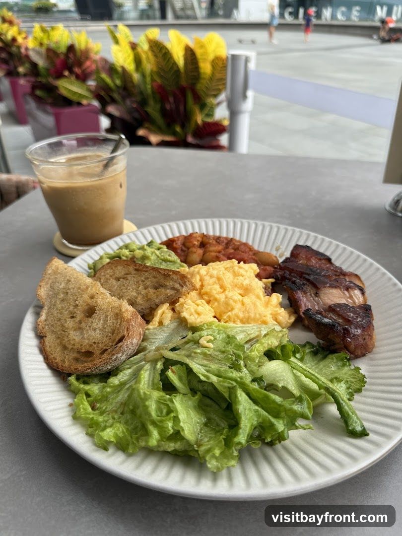 A plate of breakfast food including scrambled eggs, baked beans, avocado, salad, toast, and meat is shown on a table. An iced coffee with milk is next to the plate. In the background, there are potted plants and a blurred view of a building with the words "SCIENCE MUSEUM" visible.