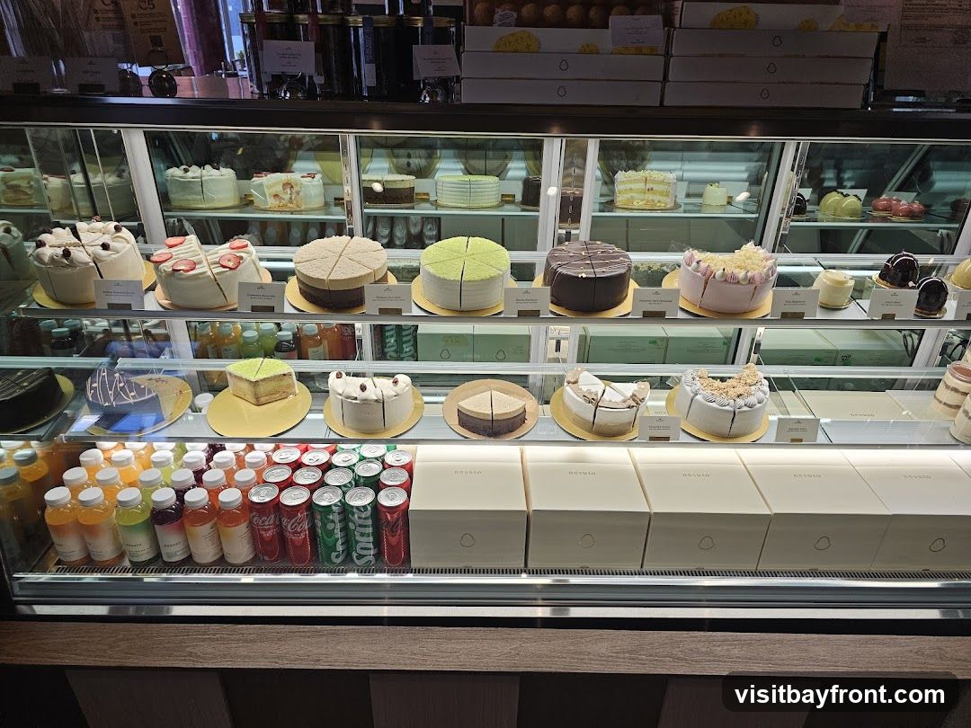 A refrigerated display case filled with various cakes, pastries, and bottled drinks. The cakes are arranged on multiple shelves, with some whole and others sliced. Below the cakes, rows of colorful bottled beverages and cans of soda are visible. The display case is well-lit, showcasing the desserts and drinks.