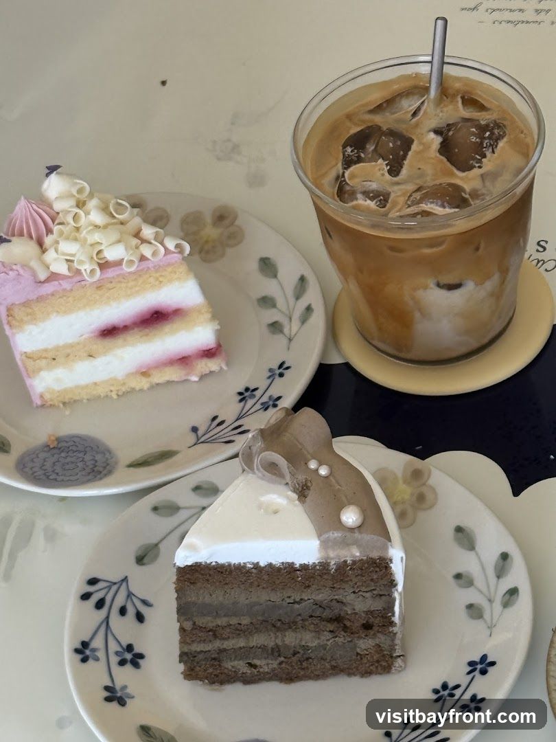 Two slices of cake and a glass of iced coffee are arranged on plates and a coaster. The cake on the left is pink and white with white chocolate curls and pink frosting. The cake on the right is brown and white with a decorative topping. The iced coffee is in a clear glass with a metal straw.