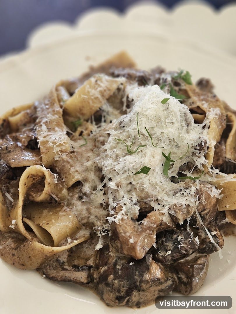 A close-up shot of a plate of pasta with a creamy mushroom sauce, topped with grated cheese and fresh herbs. The pasta appears to be pappardelle or a similar wide noodle. The mushrooms are chunky and mixed throughout the sauce.