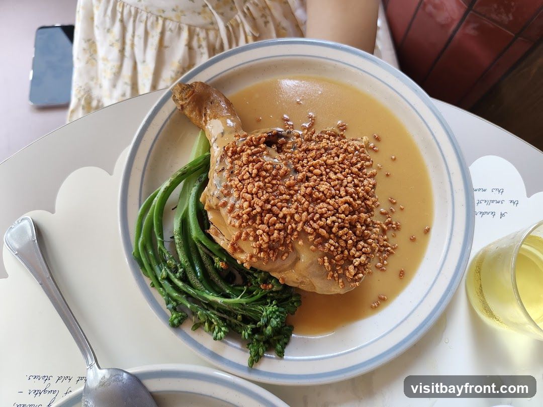 A close-up overhead shot of a plate of food. The dish features a piece of chicken leg covered in a light brown sauce and topped with crispy bits. It is served with a side of green vegetables, likely broccolini. A glass of pale yellow liquid, possibly wine or juice, is visible to the right of the plate. The setting appears to be a restaurant or cafe, with a person wearing a floral dress in the background.