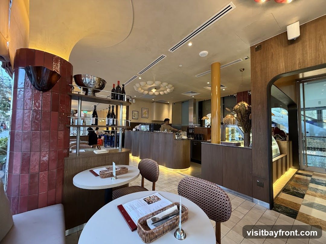 The interior of a cafe with tables and chairs. There is a counter with a barista preparing drinks. Shelves display bottles of wine and decorative items. The decor is modern with warm wood tones and tiled accents.