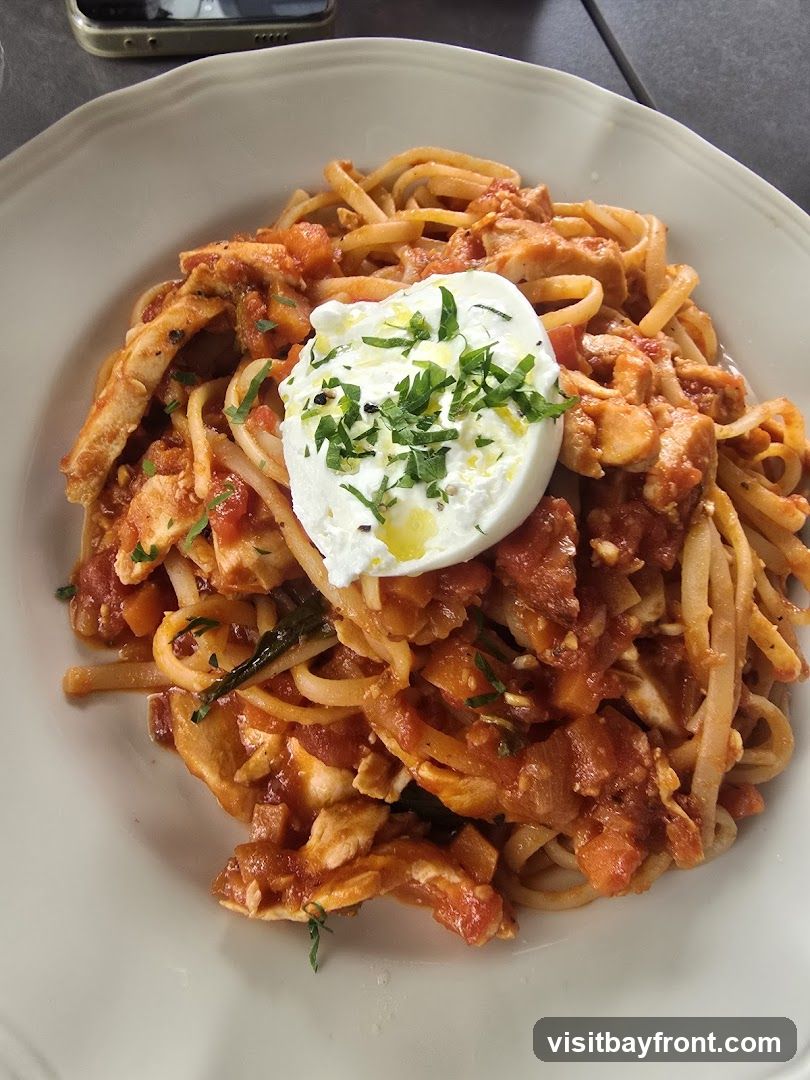 A close-up shot of a plate of pasta, likely linguine, tossed in a rich tomato sauce with shredded chicken. The dish is topped with a dollop of fresh cheese, possibly burrata or mozzarella, and garnished with chopped parsley and a drizzle of olive oil. The pasta appears to be al dente, and the sauce is chunky with visible pieces of tomato and chicken. The overall presentation suggests a hearty and flavorful meal.