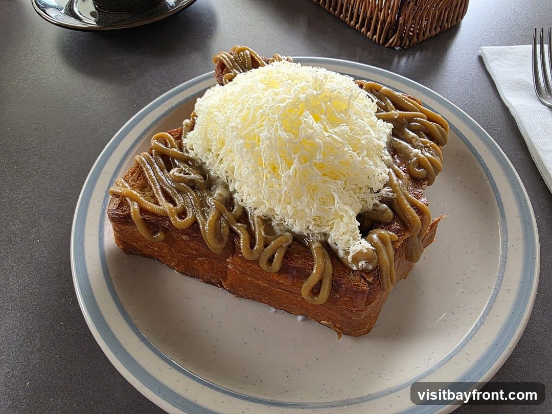 A close-up shot of a thick slice of toasted bread, possibly brioche or a similar rich pastry, served on a plate with a blue and white striped rim. The toast is generously drizzled with a thick, caramel-colored sauce and topped with a mound of finely grated white cheese, resembling shredded mozzarella or a similar mild cheese. The texture of the toast appears flaky and layered. In the background, a dark mug and saucer are visible, along with a woven basket and a white napkin with a fork.