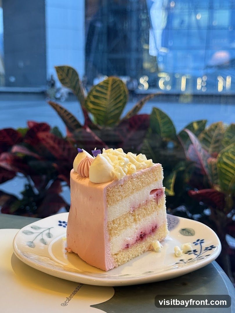 A slice of pink cake with white frosting and berry filling sits on a decorative plate. The cake has multiple layers of sponge and cream, topped with white chocolate shavings and pink frosting swirls. In the background, lush green and red plants are visible through a window, with blurred city lights and building reflections. A small watermark on the plate reads '@cafenestuto.s'.