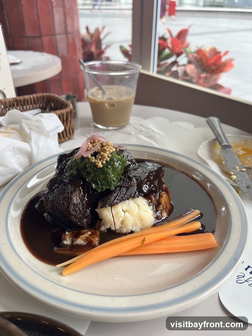 A plate of braised short ribs served with mashed potatoes, carrots, and a topping of what appears to be gremolata and pickled onions. A glass of iced coffee and a basket with napkins are visible in the background.