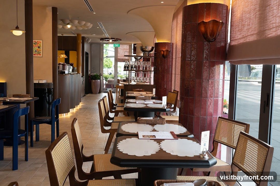 The interior of a restaurant with tables and chairs set up for dining. The decor features warm lighting, wooden furniture, and decorative pillars. An exit sign is visible in the background.