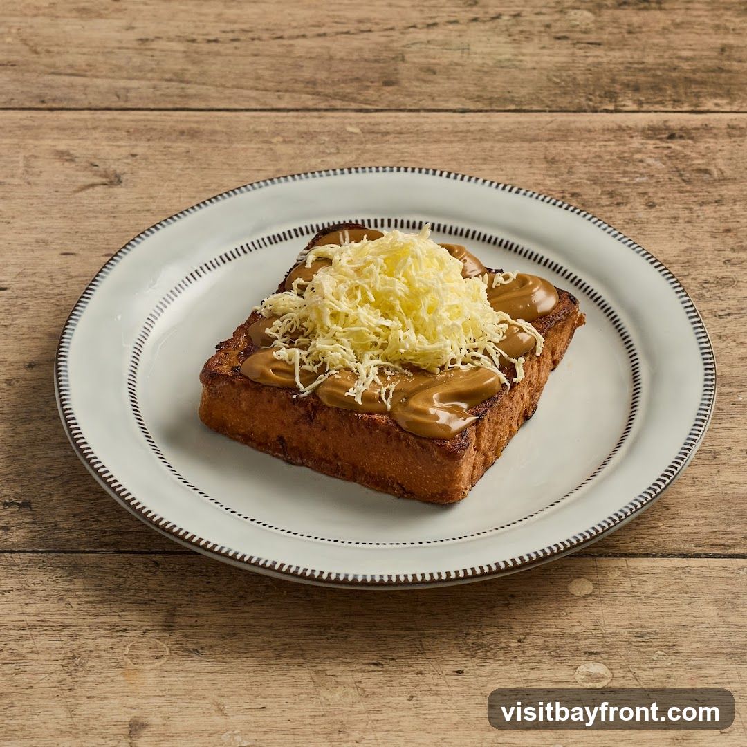A slice of thick-cut French toast, golden brown and crispy, is served on a white plate with a decorative rim. The toast is generously topped with a swirl of smooth caramel sauce and a mound of shredded cheese. The plate rests on a rustic wooden surface.