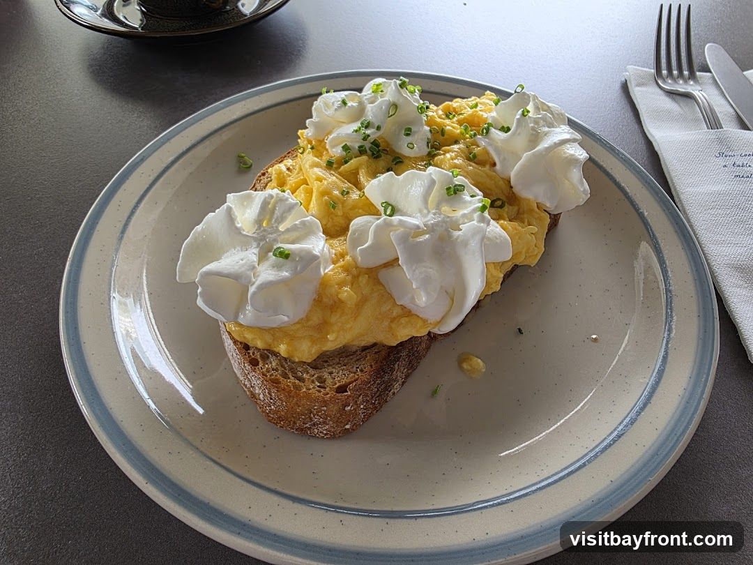 A close-up shot of scrambled eggs served on a slice of toasted bread, topped with dollops of whipped cream and chives. The dish is presented on a plate with a blue and white rim, set on a dark grey surface. A napkin with cutlery and some text is visible to the right.