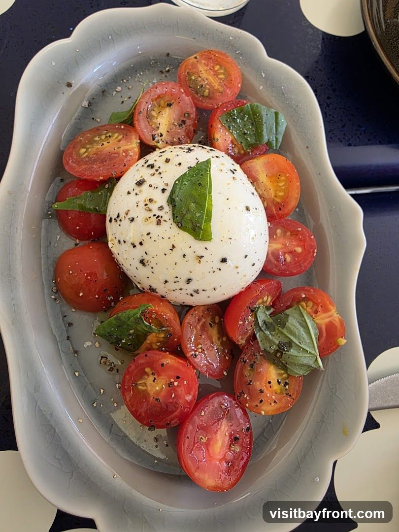 A close-up overhead shot of a Caprese salad served in a light blue, scalloped dish. The salad features a large ball of fresh mozzarella cheese in the center, surrounded by halved cherry tomatoes. Fresh basil leaves are scattered throughout, and the dish is seasoned with black pepper and salt. The lighting is bright, highlighting the vibrant colors of the ingredients.