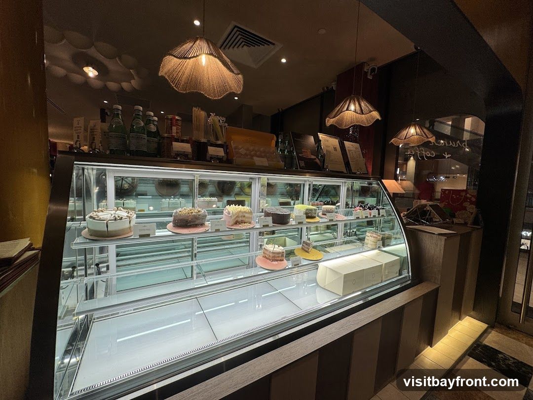 A refrigerated display case filled with various cakes and pastries. The case is well-lit, showcasing the desserts on multiple shelves. Above the case, bottles of San Pellegrino water and other items are visible on a shelf. Decorative pendant lights hang from the ceiling, casting a warm glow. The overall ambiance suggests a bakery or cafe.