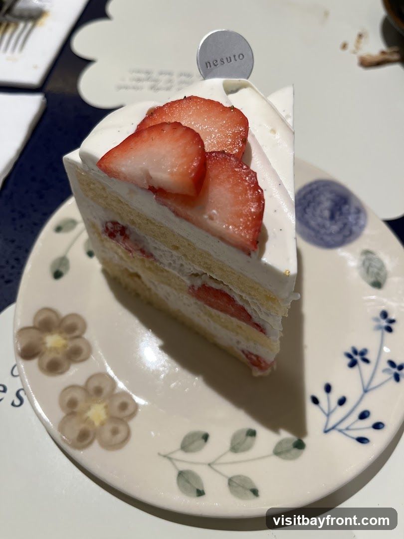 A slice of strawberry shortcake with layers of sponge cake, cream, and strawberries, topped with whipped cream and fresh strawberry slices. A small silver tag with the word "nesuto" is inserted into the cake. The cake is served on a decorative plate with floral patterns.