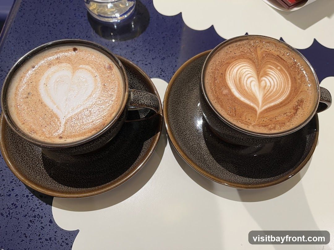 Two cups of coffee with latte art in the shape of a heart are on saucers. The cups and saucers are dark brown with a speckled texture. The coffee on the left has a simpler heart shape, while the coffee on the right has a more intricate latte art design. The cups are placed on a table with a blue and white patterned surface.