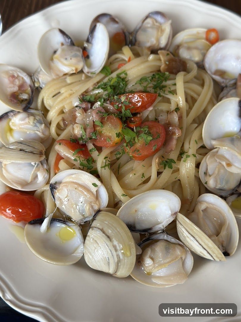 A close-up shot of a plate of linguine pasta with clams, cherry tomatoes, and parsley. The pasta is coated in a light sauce, and the clams are arranged around the edges of the dish.