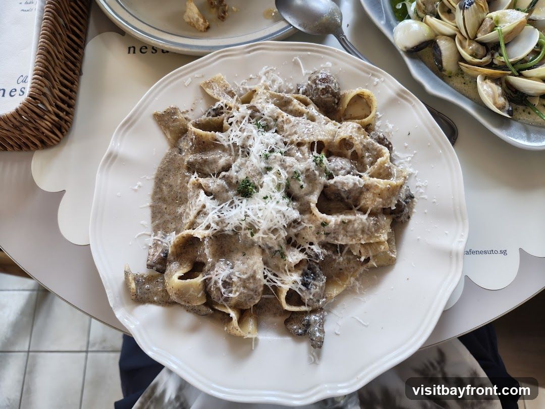 A close-up overhead view of a plate of pappardelle pasta with a creamy mushroom sauce and grated cheese. To the right, a dish of clams with onions and greens is visible. A wicker basket and a menu are partially visible on the left. The text 'cafenesuto.sg' is visible on a placemat.