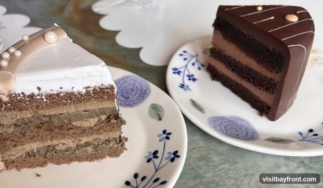 A close-up shot of two slices of cake on decorative plates. The slice on the left is a layered cake with white frosting and a mocha-colored drizzle, adorned with pearls. The slice on the right is a rich chocolate cake with chocolate frosting and a glossy chocolate glaze, decorated with white lines and gold accents. Both cakes are presented on white plates with blue floral and green leaf designs. A white tiered cake stand is visible in the background.